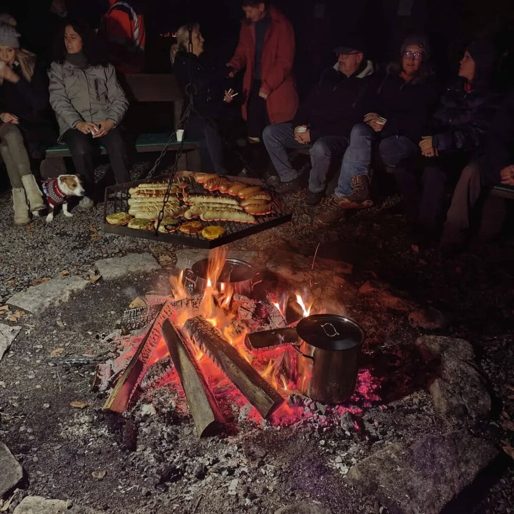 Schwedenfeuer mit Grillgut und Menschen am Lagerfeuer bei Nacht, gemütliches Beisammensein im Freien.